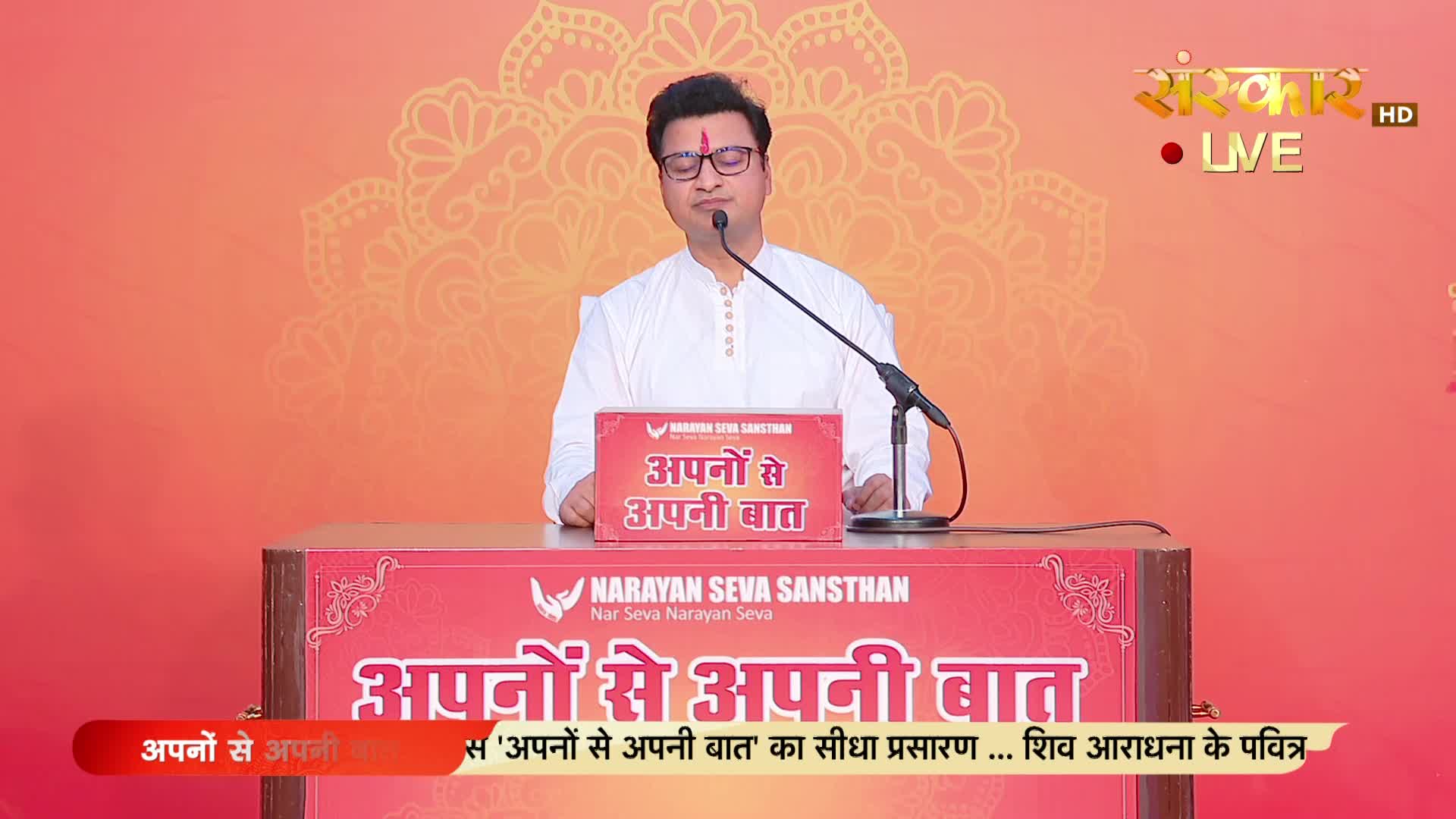 A man in glasses and a white kurta stands behind a podium, speaking into a microphone. The Sanskar TV logo and the words "LIVE" are visible in the top right corner, and the backdrop is a warm orange.
