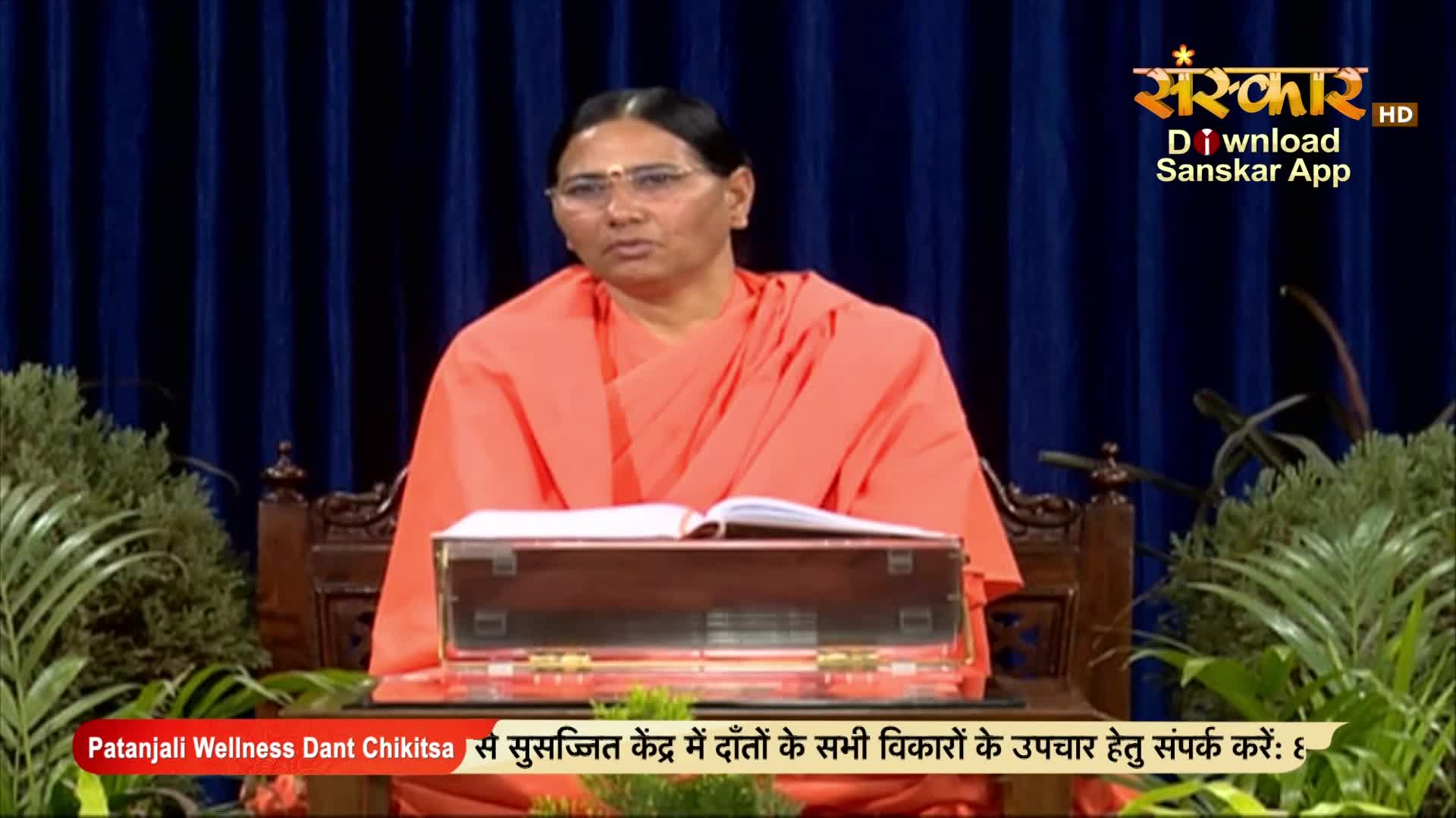 A woman in an orange robe sits behind a wooden desk, a book open in front of her. The Sanskar TV logo and Hindi text are visible in the background.
