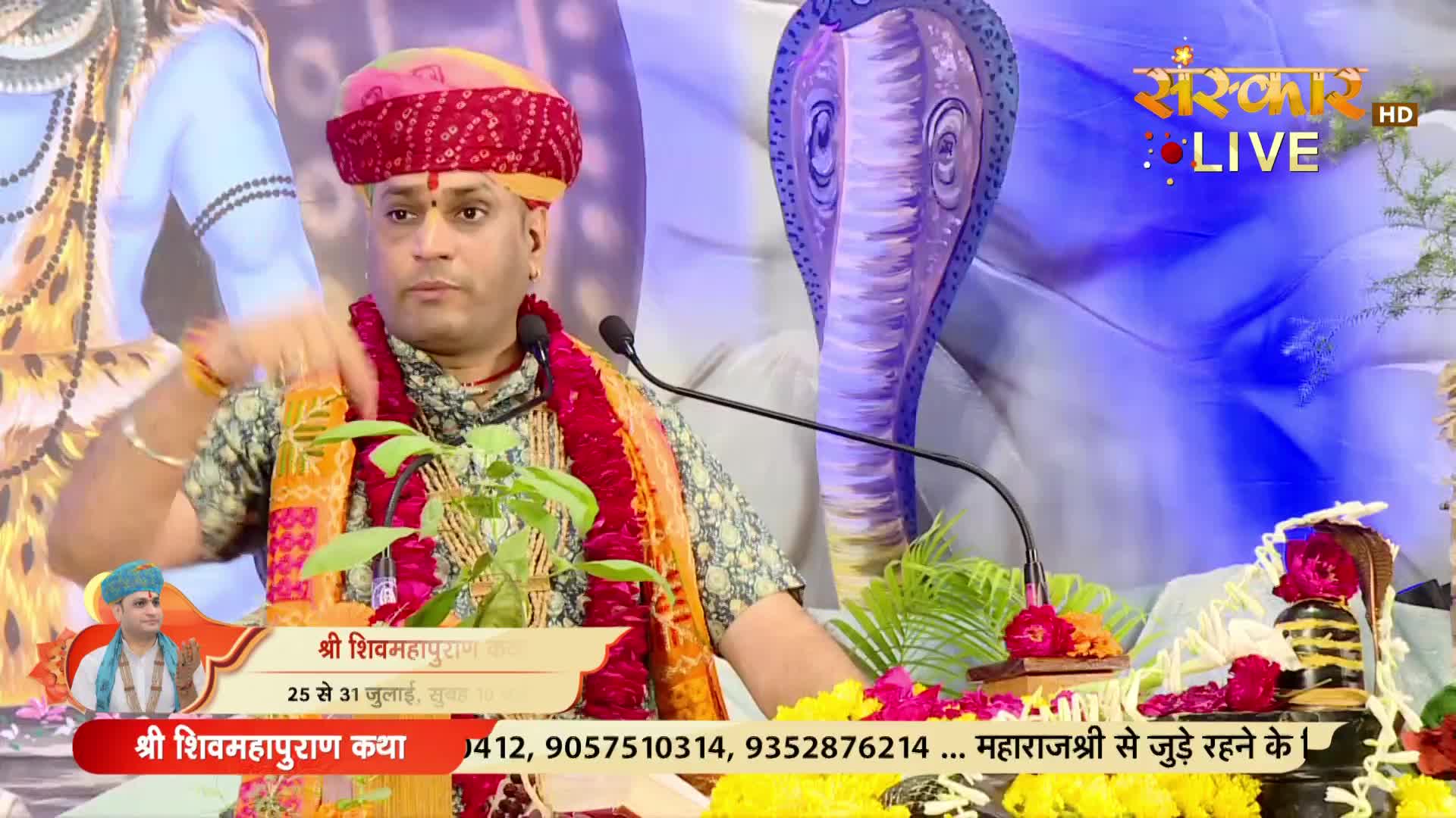 A man in a turban and garland gestures with his hand while speaking into a microphone. The Sanskar TV logo is visible, and the bottom banner indicates the event is "Shri Shivmahapuran Katha" in India.
