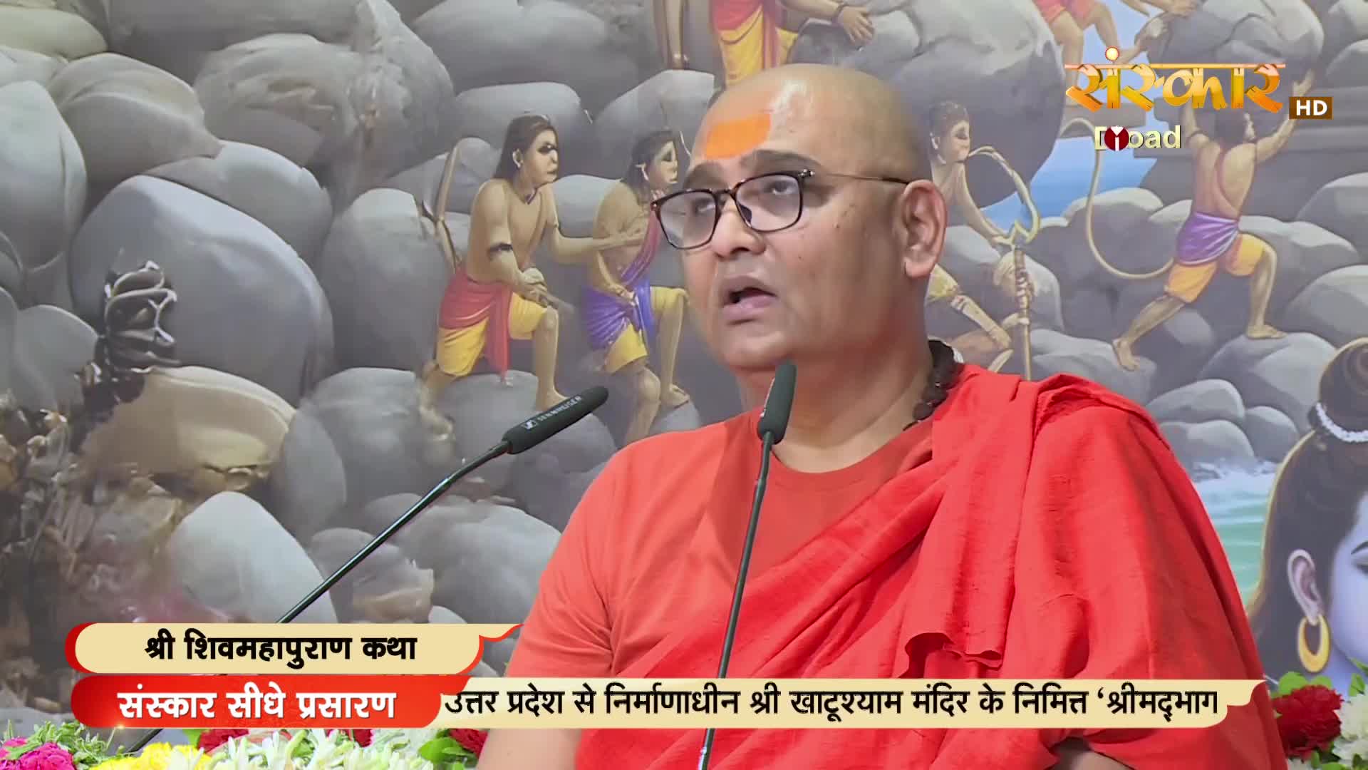A man in an orange robe speaks earnestly into a microphone, his face framed by glasses and a bald head with a saffron mark. Behind him, a backdrop depicts figures and large stones, likely part of a religious broadcast from India on Sanskar TV.
