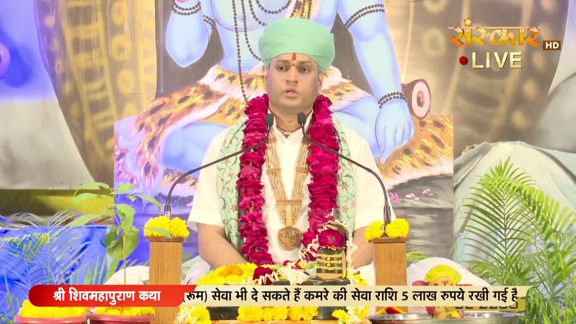A man in a green turban and garland speaks into two microphones, likely on Sanskar TV, with a backdrop depicting a deity. Below, a banner displays text in Hindi, and the scene is framed by floral arrangements.
