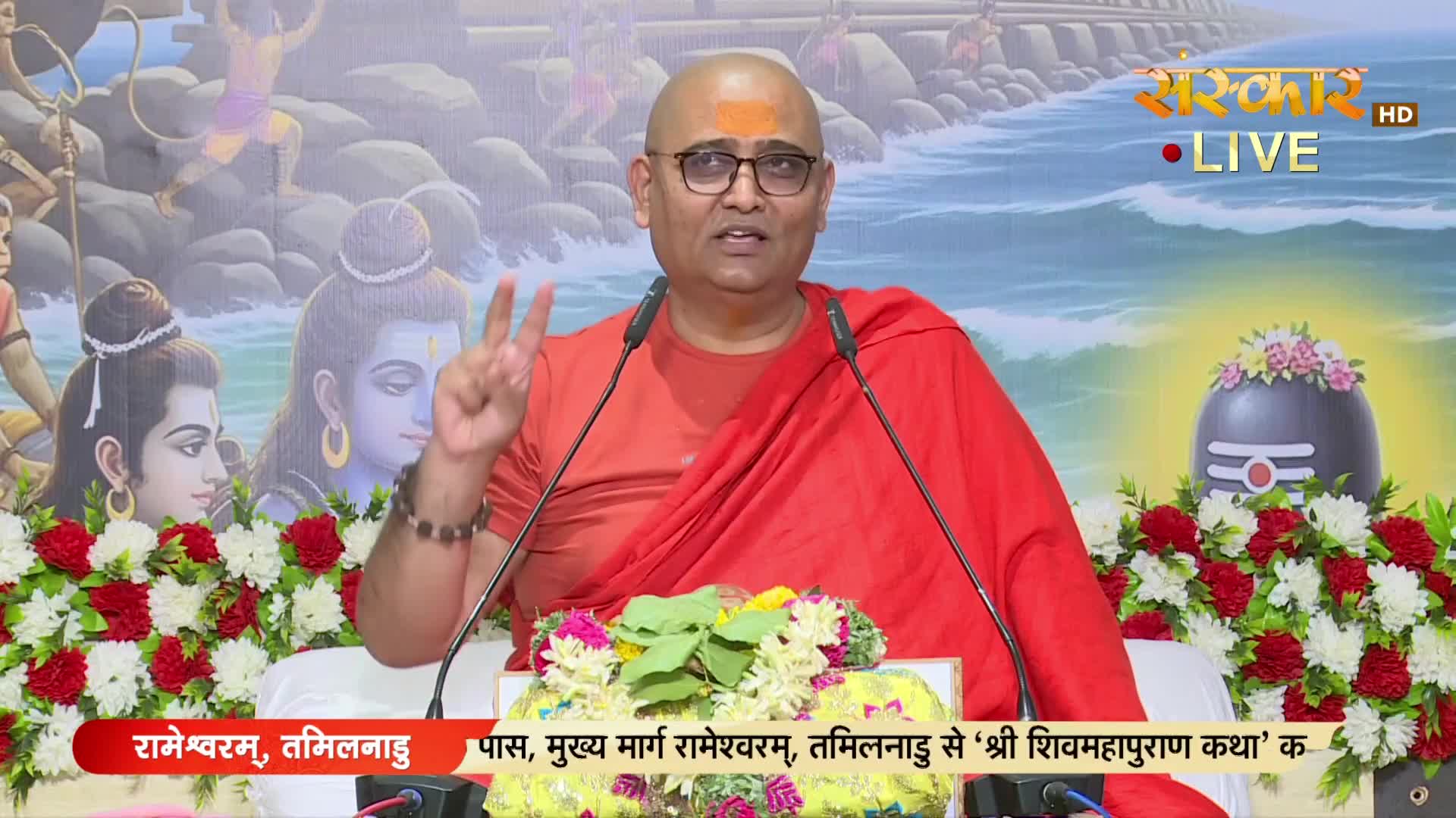 A man in an orange robe gestures with two fingers raised, speaking into a microphone. The backdrop features religious imagery and a Sanskar TV logo, with text in Hindi at the bottom.

