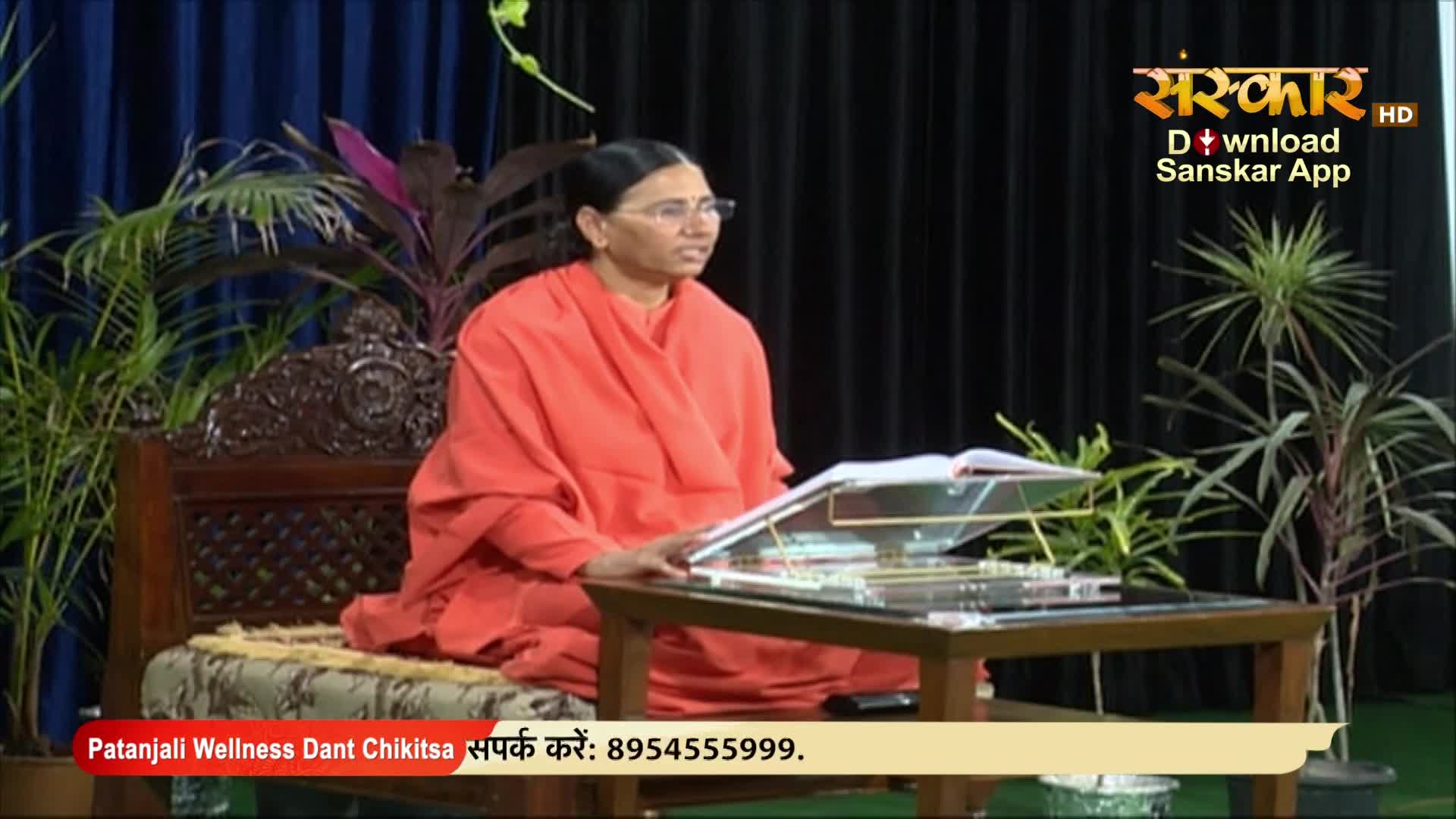 A woman in an orange robe sits at a table, speaking into the camera on Sanskar TV. She's surrounded by plants, with a book open in front of her.
