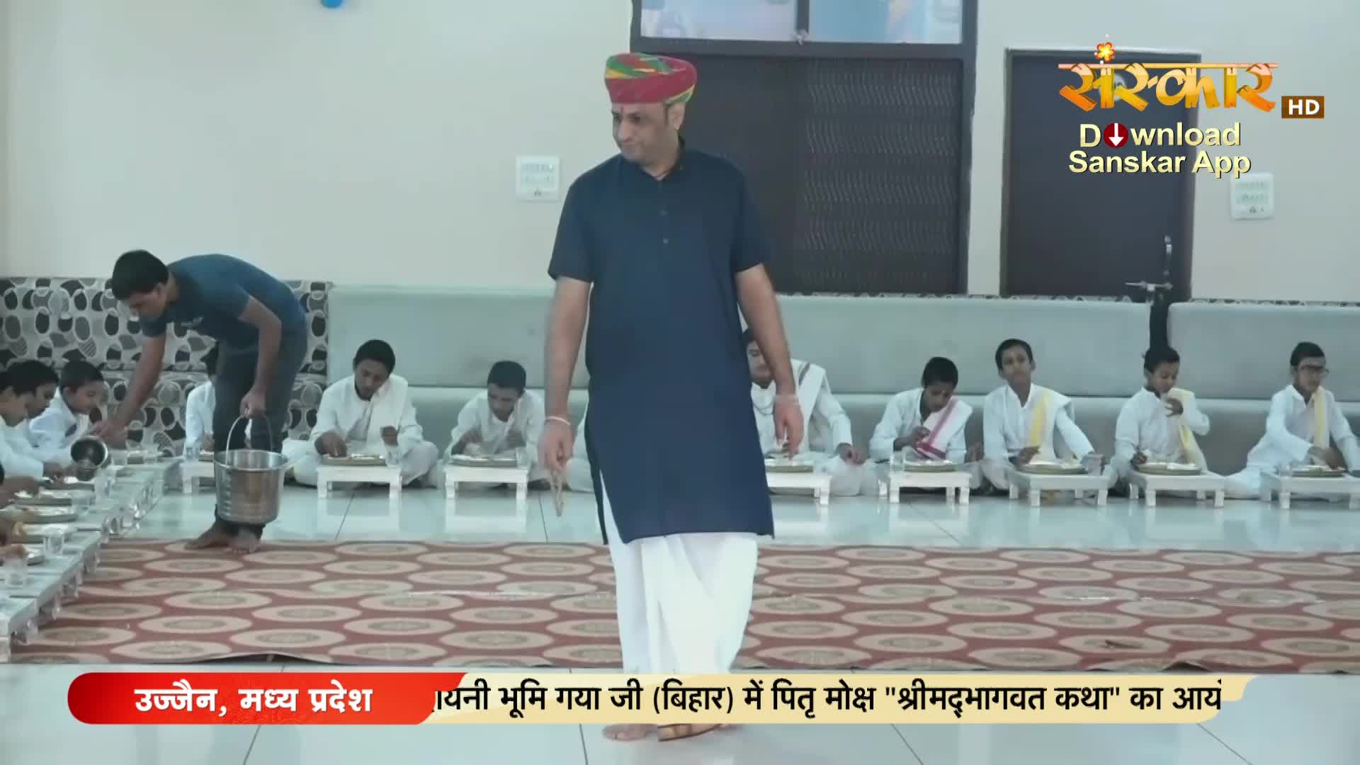 A man in a dark kurta walks past the camera, while a younger man pours from a metal bucket. Several children in white robes sit on the floor, eating from small plates.
