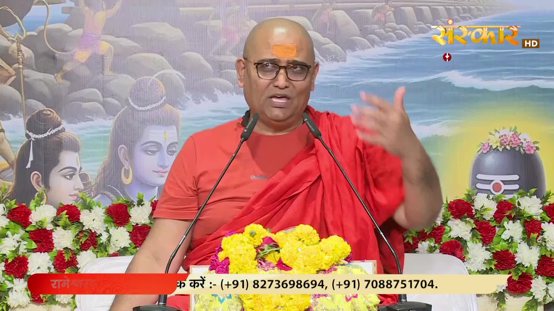 A man in a red robe gestures with his right hand as he speaks into two microphones. Behind him, a backdrop shows religious figures and the Sanskar TV logo.
