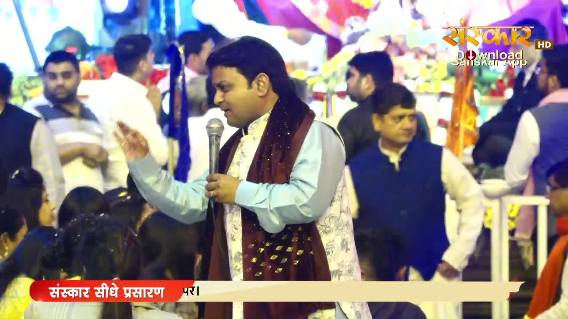 A man in a patterned jacket and maroon scarf speaks into a microphone, gesturing with his right hand. The scene is being broadcast by Sanskar TV, with a crowd gathered in the background.
