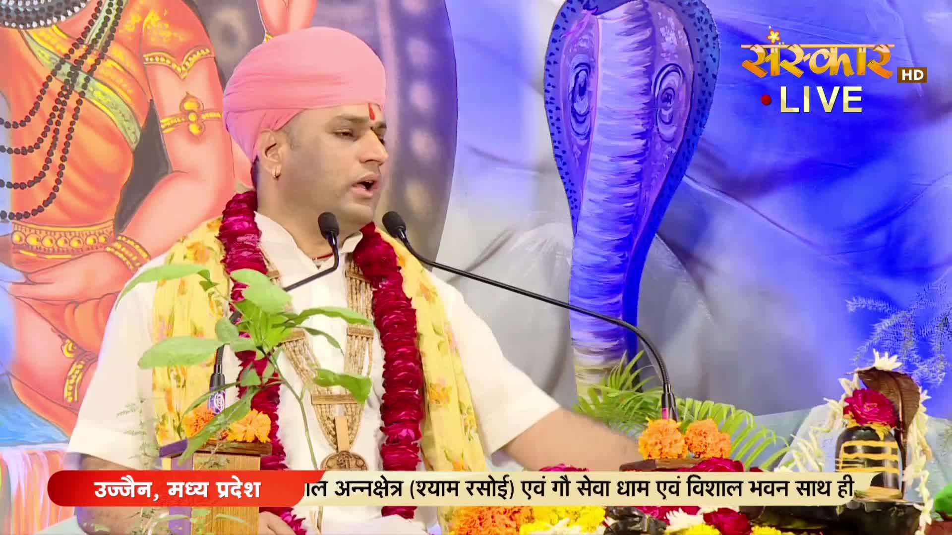 A man in a pink turban and white clothing speaks into a microphone, a garland draped around his neck. The Sanskar TV logo is visible, and the backdrop features a large, stylized cobra head.
