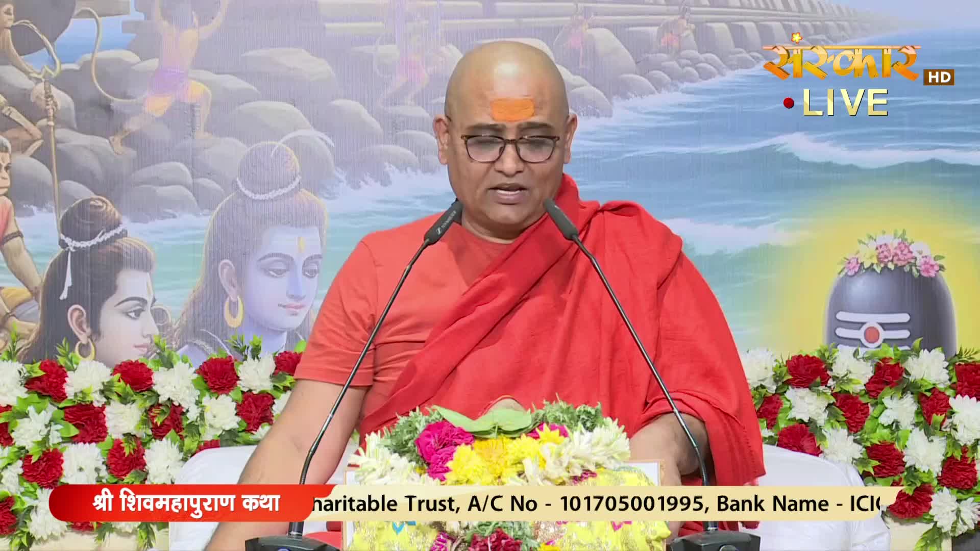 A man in a saffron robe speaks into two microphones, his brow marked with a red tilak. The scene is broadcast live on Sanskar TV, with a backdrop of religious imagery.
