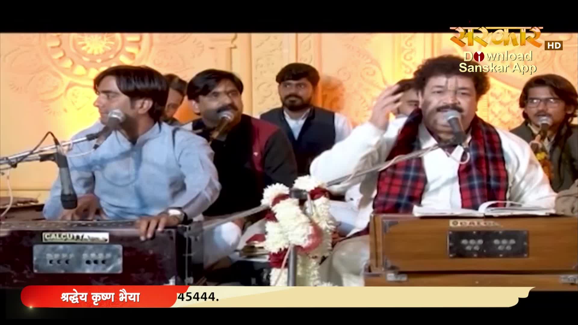 A man in a white shirt and scarf gestures emphatically while singing into a microphone. Behind him, other musicians accompany him, their hands moving over the harmoniums.
