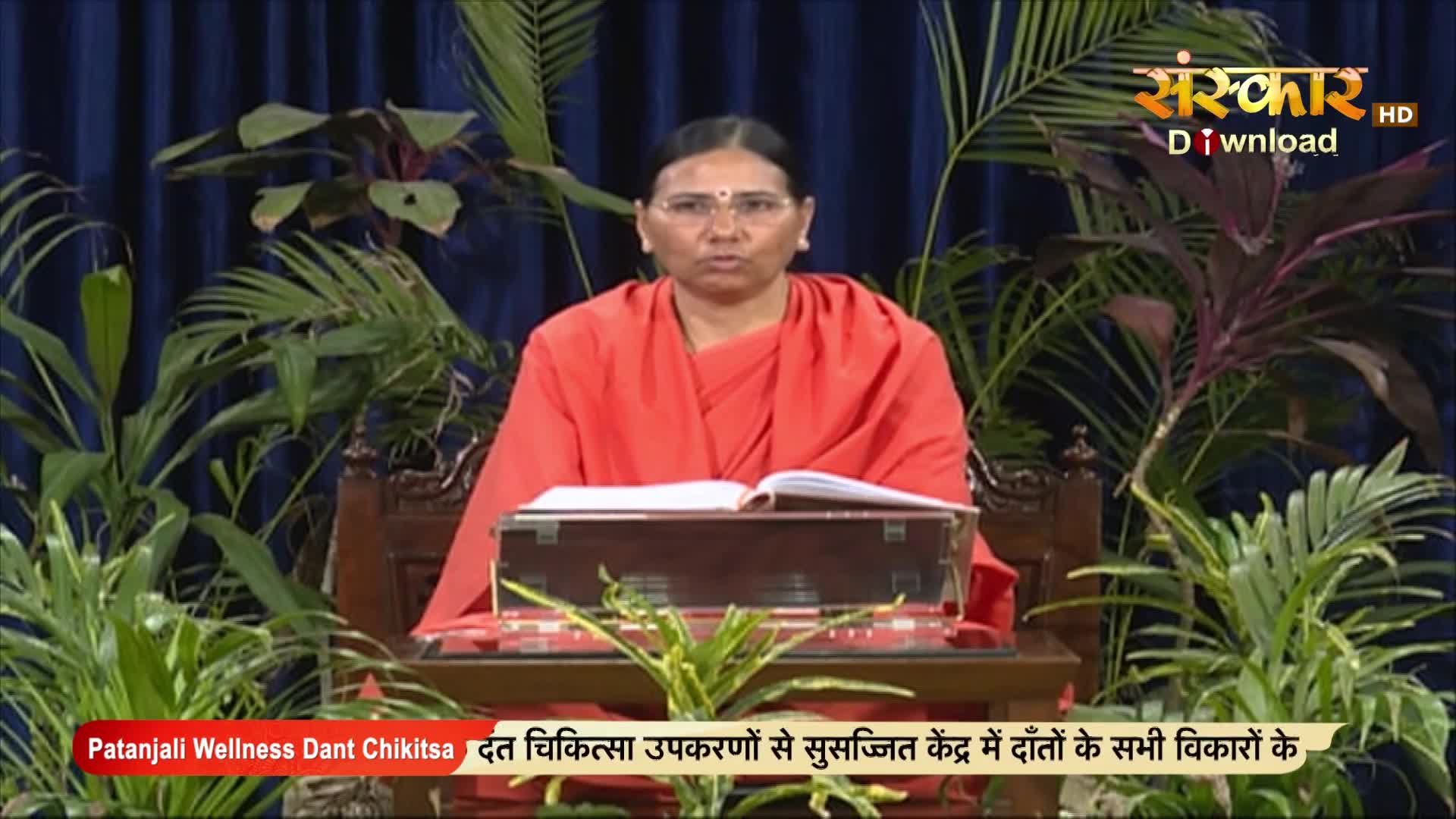 A woman in an orange robe sits at a wooden desk, a book open in front of her. The Sanskar TV logo is visible in the top right corner, with text at the bottom advertising "Patanjali Wellness Dant Chikitsa."
