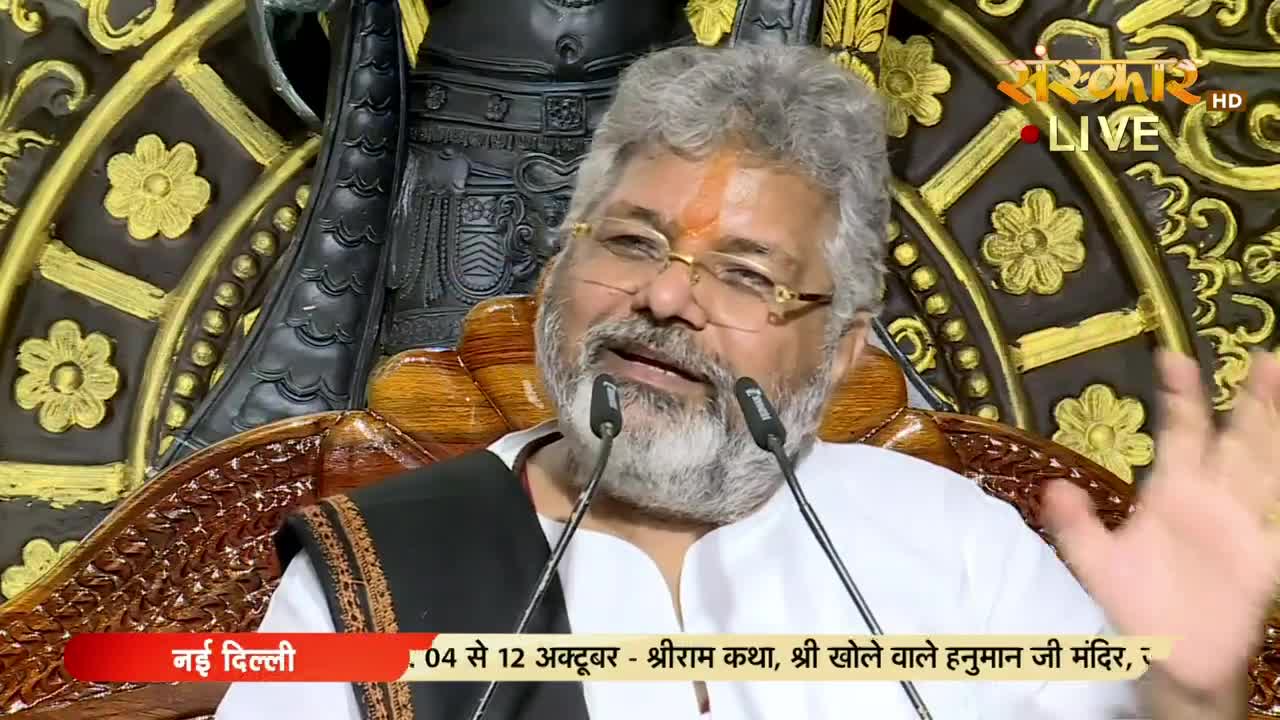 A man with a silver beard and glasses gestures with his right hand while speaking into two microphones. The Sanskar TV logo is visible in the top right corner, and a banner at the bottom indicates the event is in New Delhi.
