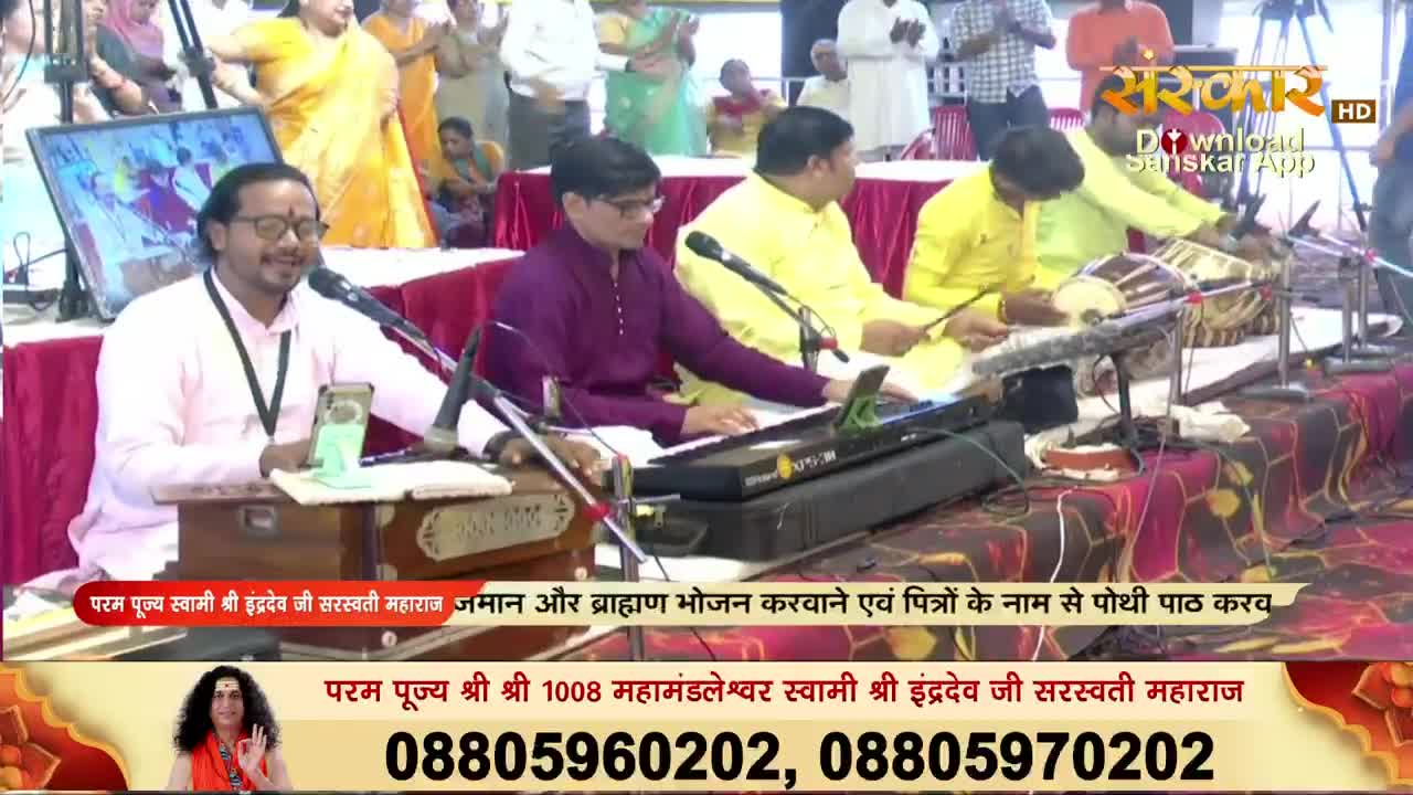 A man in a pink shirt sings into a microphone, his eyes closed, while a keyboardist and percussionists play alongside him. The scene is broadcast on Sanskar TV, with a banner promoting the event.
