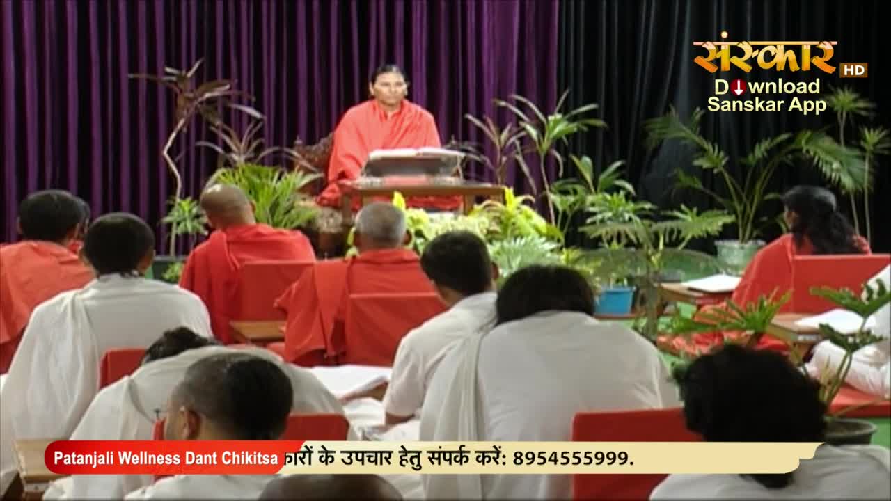 A person in an orange robe is speaking from behind a table, with a group of people in front of them, some wearing white and some wearing red. The scene is being broadcast on Sanskar TV, with a phone number for Patanjali Wellness displayed at the bottom.
