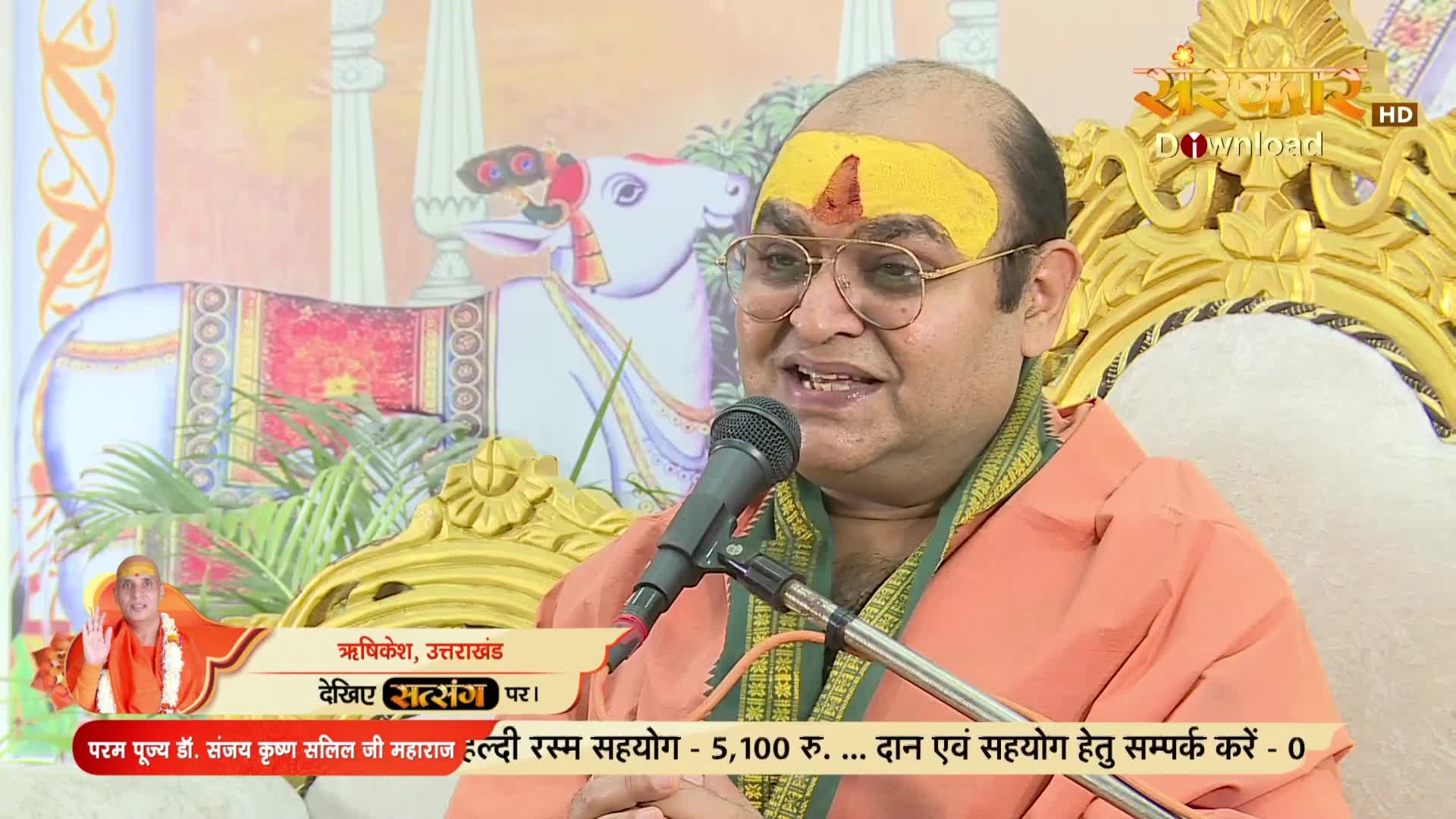 A man with a yellow mark on his forehead speaks into a microphone, his face lit by the studio lights. Behind him, a decorative cow statue sits next to a golden chair, all part of the set for a Sanskar TV broadcast.
