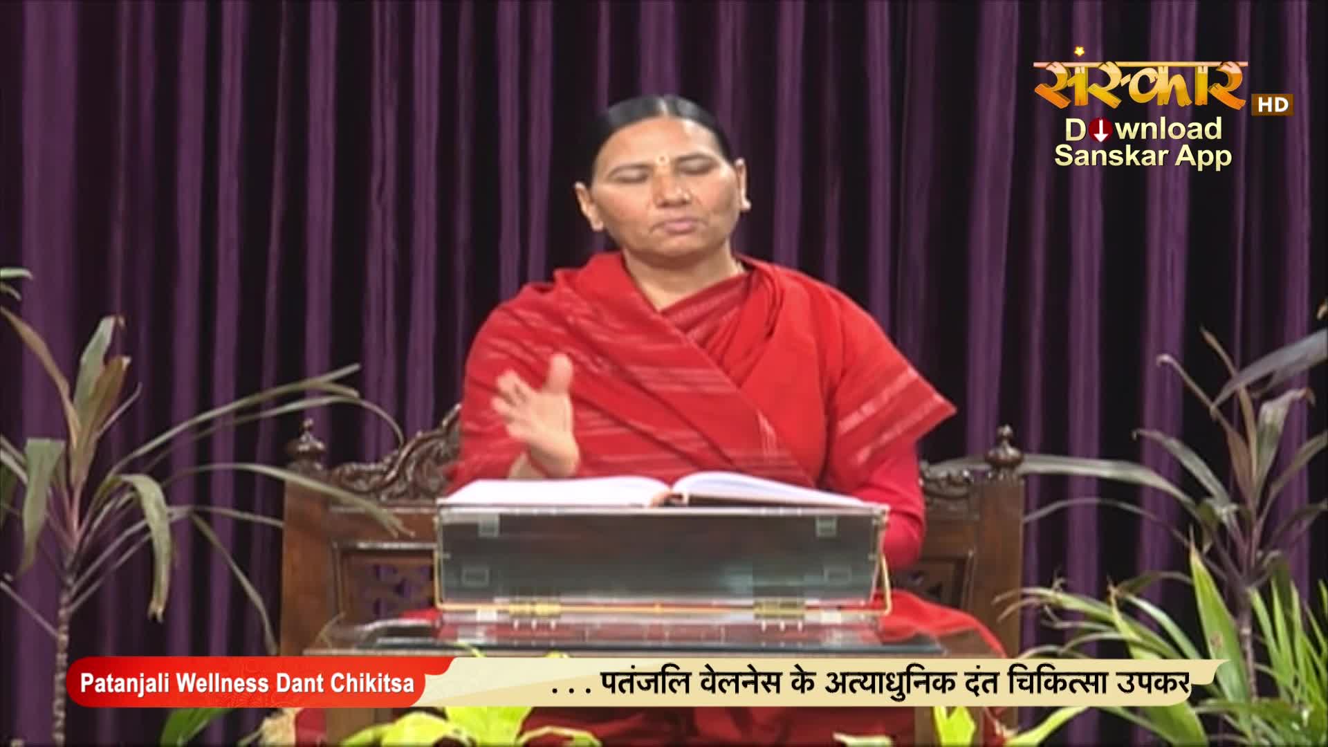 A woman in a red shawl gestures with her right hand while speaking, seated behind a small table with an open book. The Sanskar TV logo is visible, and the program is about Patanjali Wellness.
