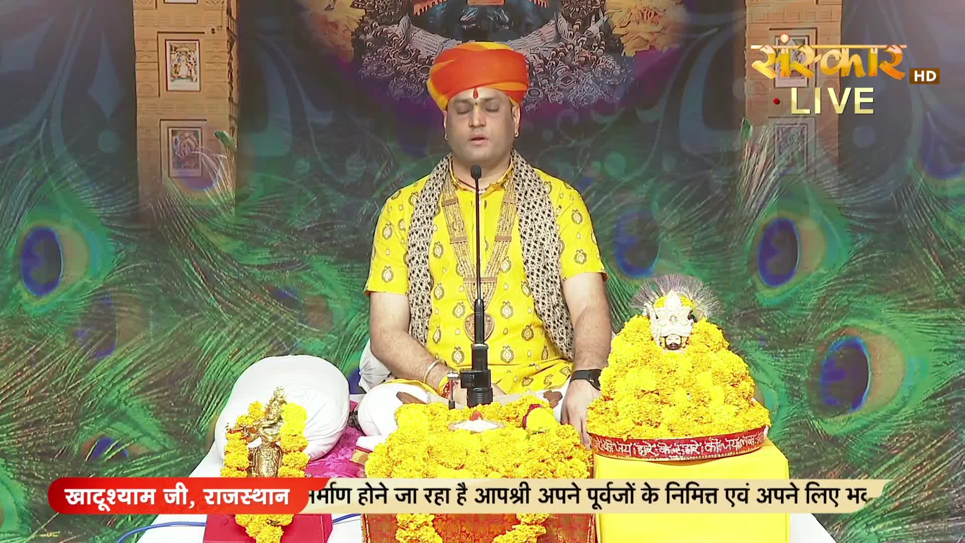 A man in a yellow shirt and orange turban sits with his eyes closed, speaking into a microphone. The backdrop is decorated with peacock feather patterns, and the Sanskar TV logo is visible in the corner.
