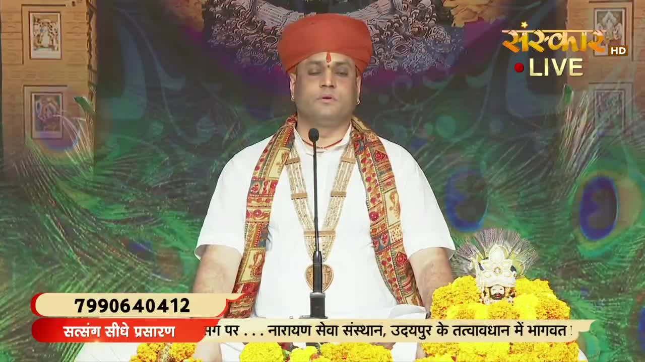 A man in a turban and white clothing speaks into a microphone, his face illuminated by the studio lights. The scene is broadcast live on Sanskar TV, with text in Hindi at the bottom of the screen.
