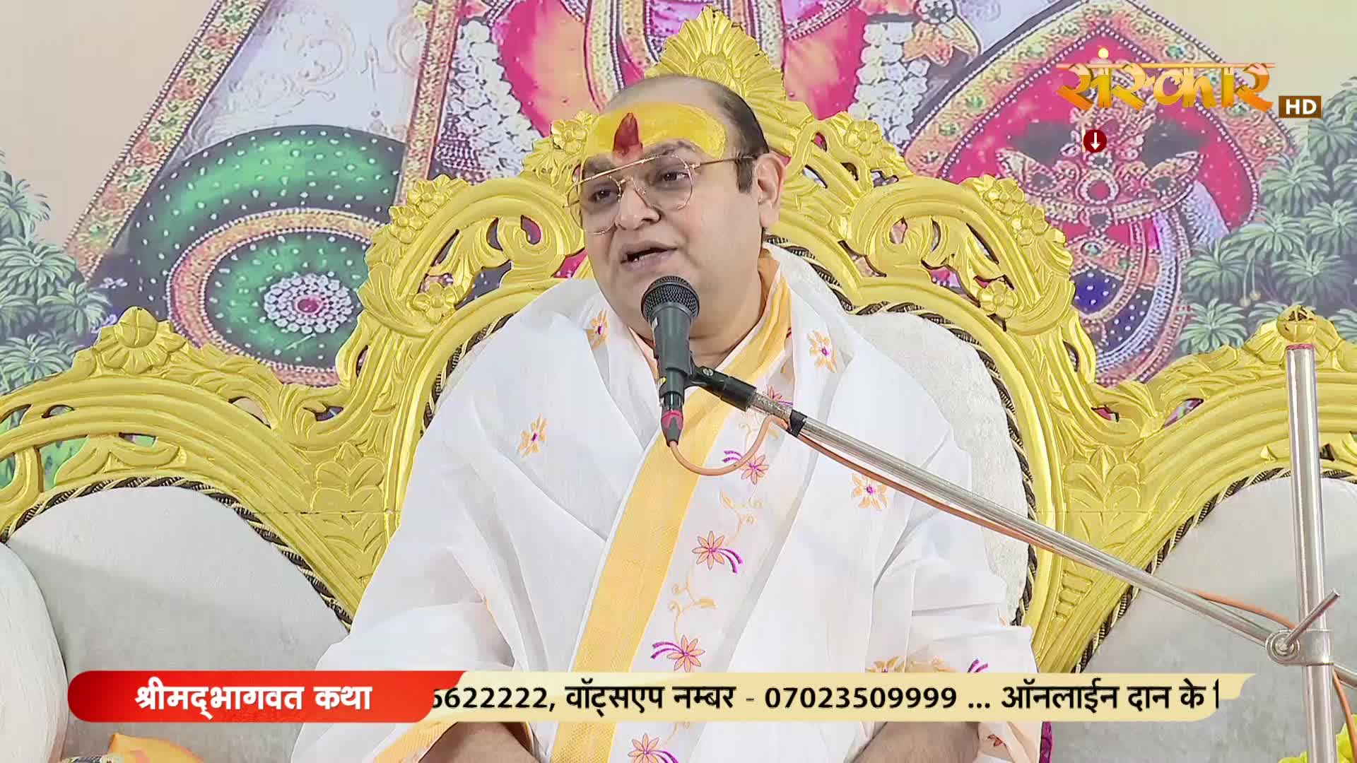 A man in a white robe speaks into a microphone, his face illuminated on a set decorated with gold detailing. The Sanskar TV logo is visible in the top right corner, and text at the bottom indicates a live broadcast from India.
