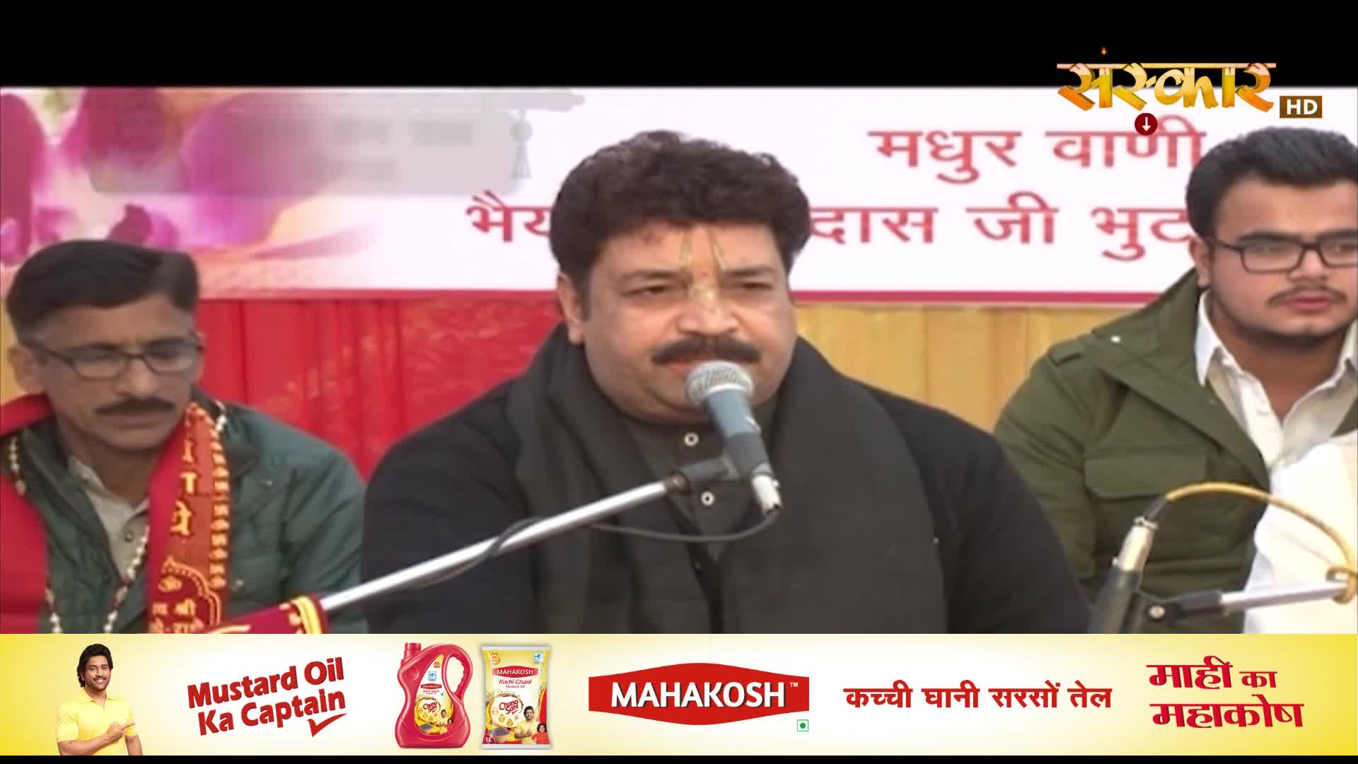 A man with a mustache speaks into a microphone, likely on Sanskar TV, while two others sit beside him. The backdrop is a simple banner, and the bottom of the screen displays an advertisement for mustard oil.
