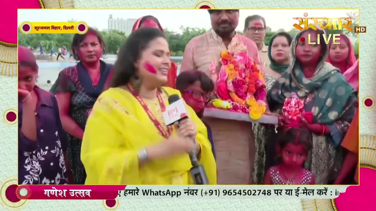 A woman in a yellow outfit, her face marked with color, speaks into a microphone, while a group of people, some with painted faces, hold a Ganesh idol.  The scene is being broadcast live on Sanskar TV during what appears to be a Ganesh Utsav celebration in Delhi.
