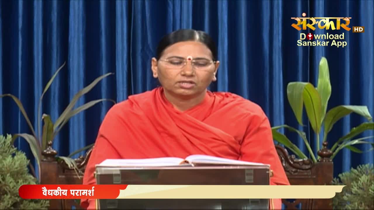 A woman in an orange robe sits behind a wooden lectern, speaking with a book open in front of her. The Sanskar TV logo is visible in the upper right corner, and a banner at the bottom reads "Vaidkiya Paramarsh."
