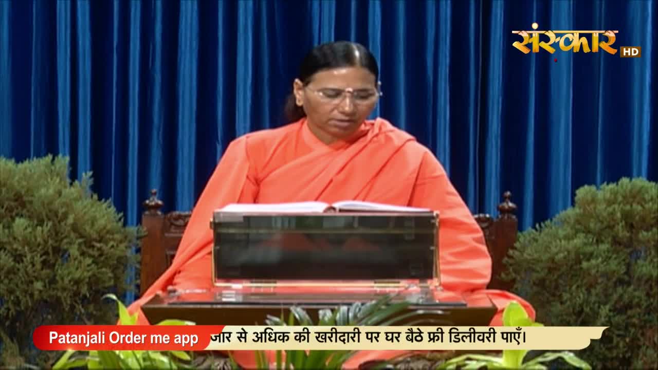 A woman in orange robes sits at a desk, reading from an open book. The Sanskar HD logo and a Patanjali ad are visible on the screen.
