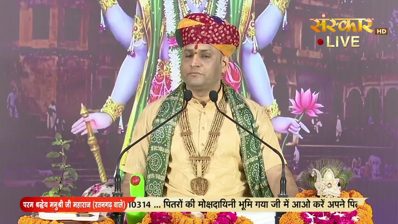 A man in a turban and shawl speaks into two microphones, a gold pendant hanging at his chest. The Sanskar TV logo shines in the corner, and a colorful backdrop of a deity is behind him.
