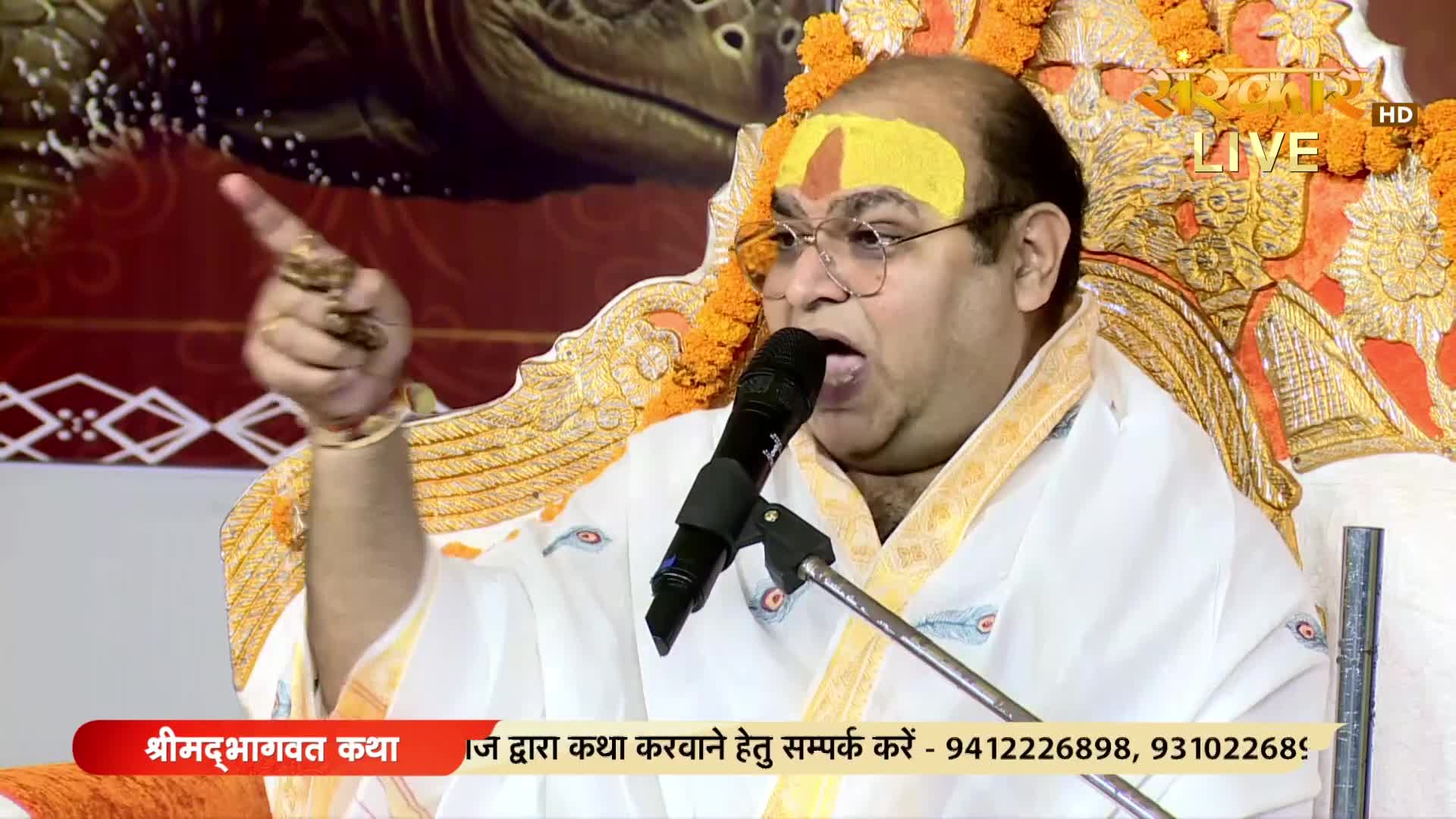 A man in a white robe and glasses gestures emphatically while speaking into a microphone, his face animated. The Sanskar TV logo is visible, and a banner at the bottom advertises a religious event in India.
