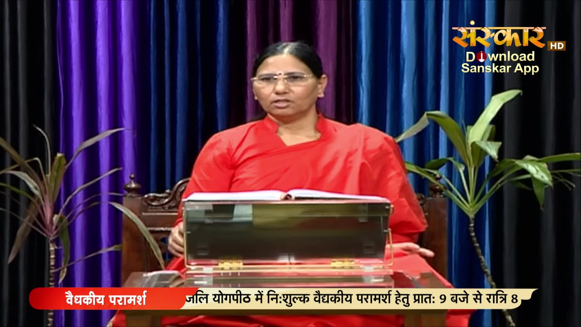 A woman in a red robe sits behind a desk, speaking directly to the camera. The Sanskar TV logo and Hindi text are visible in the background.
