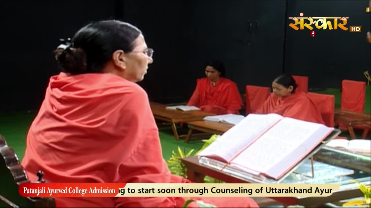 A woman in a bright orange robe sits facing away, a large book open on a stand in front of her. Across the room, two other women in similar robes sit at tables, books open before them, as the Sanskar TV logo glows in the corner.

