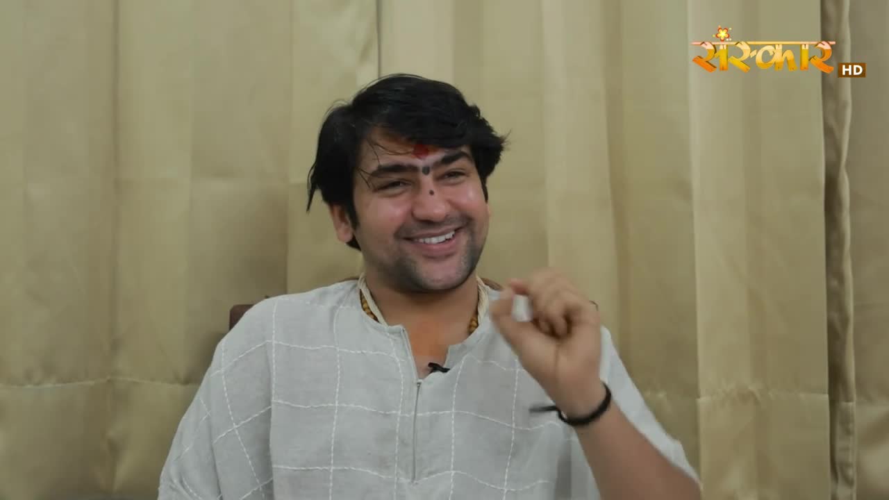 A man in a light-colored shirt, with a red mark on his forehead, smiles and gestures with his right hand. Behind him, the Sanskar HD logo is visible, suggesting a broadcast from India.
