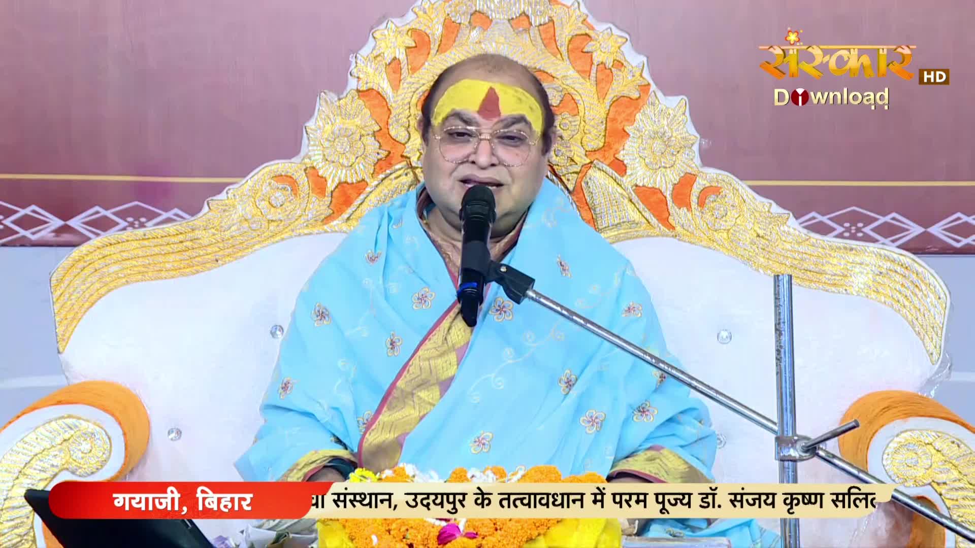 A man in glasses and a light blue shawl speaks into a microphone, seated on an ornate chair. The Sanskar TV logo is visible in the top right corner, and a banner at the bottom identifies the location as Gaya, Bihar.

