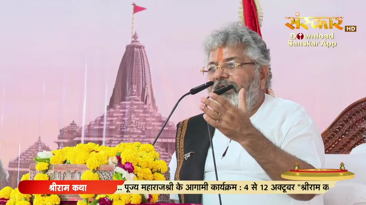 A man with a grey beard and glasses speaks into a microphone, gesturing with his right hand. The scene is set in India, likely for a broadcast on Sanskar TV, with a temple visible in the background.
