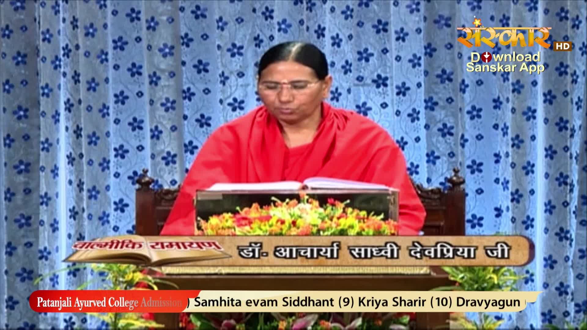 A woman in a red robe sits behind a desk, reading from a book on Sanskar TV. The set is decorated with flowers and a backdrop of blue floral curtains.
