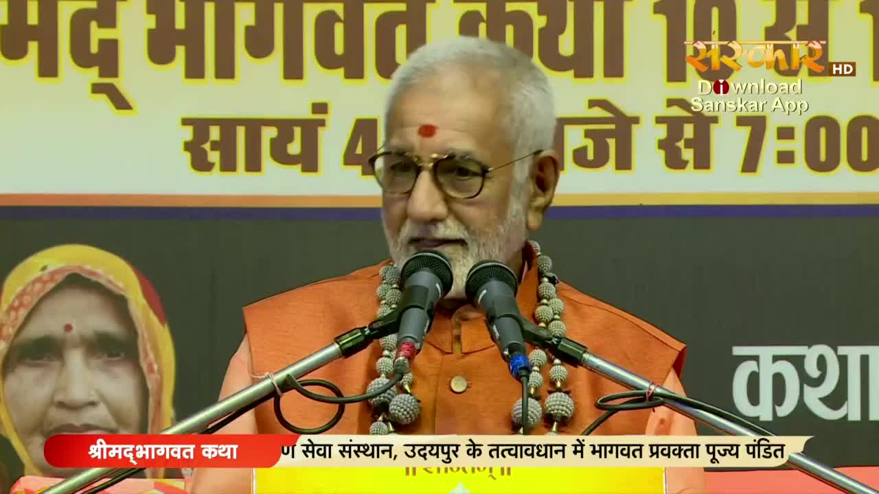A man in orange robes speaks into two microphones, his face framed by glasses and a red dot on his forehead. The Sanskar TV logo is visible in the upper right corner, with Hindi text above and below the speaker.

