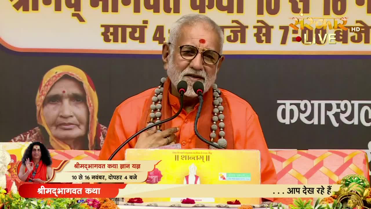 A man in orange robes speaks into two microphones, his face illuminated by the stage lights. Behind him, a woman's portrait is displayed, and the Sanskar TV logo indicates a live broadcast from India.
