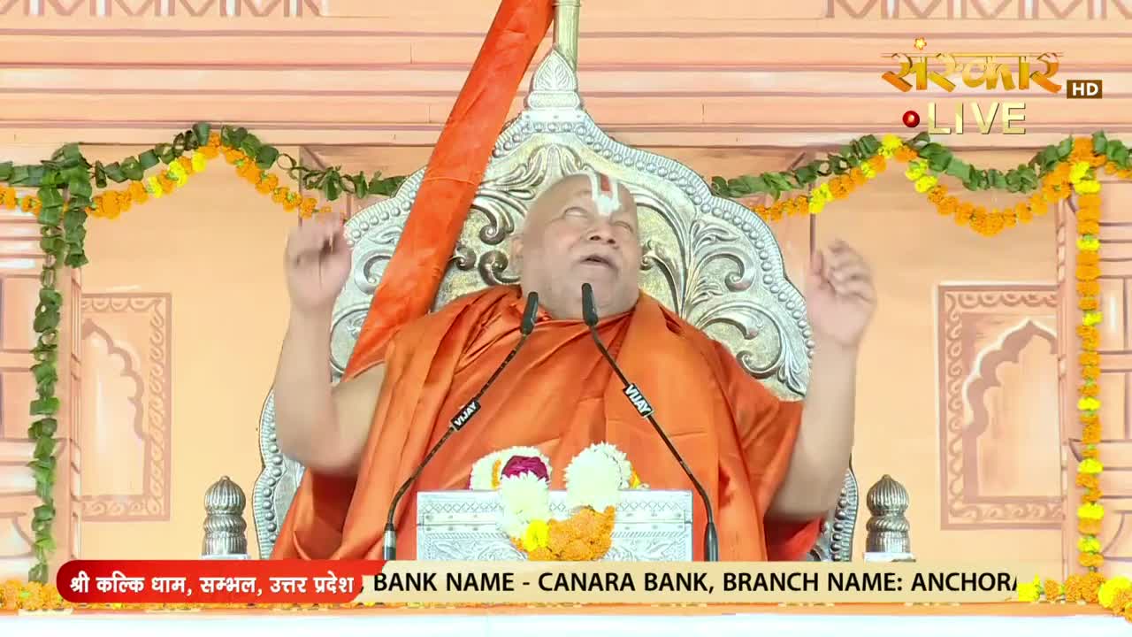 A robed man gestures with both hands as he speaks into two microphones. The scene is being broadcast live on Sanskar TV, with the location details and bank information displayed below.
A robed man gestures with both hands as he speaks into two microphones. The scene is being broadcast live on Sanskar TV, with the location details and bank information displayed below.