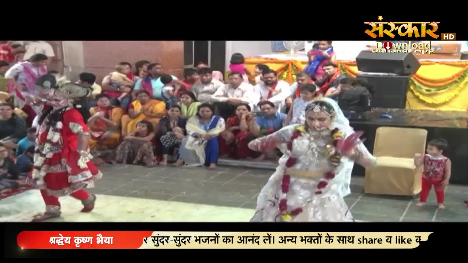 A dancer in a red costume moves across the floor, while another in white performs nearby. The audience, seated on the ground, watches the performance, which appears to be broadcast on Sanskar TV.
A dancer in a red costume moves across the floor, while another in white performs nearby. The audience, seated on the ground, watches the performance, which appears to be broadcast on Sanskar TV.