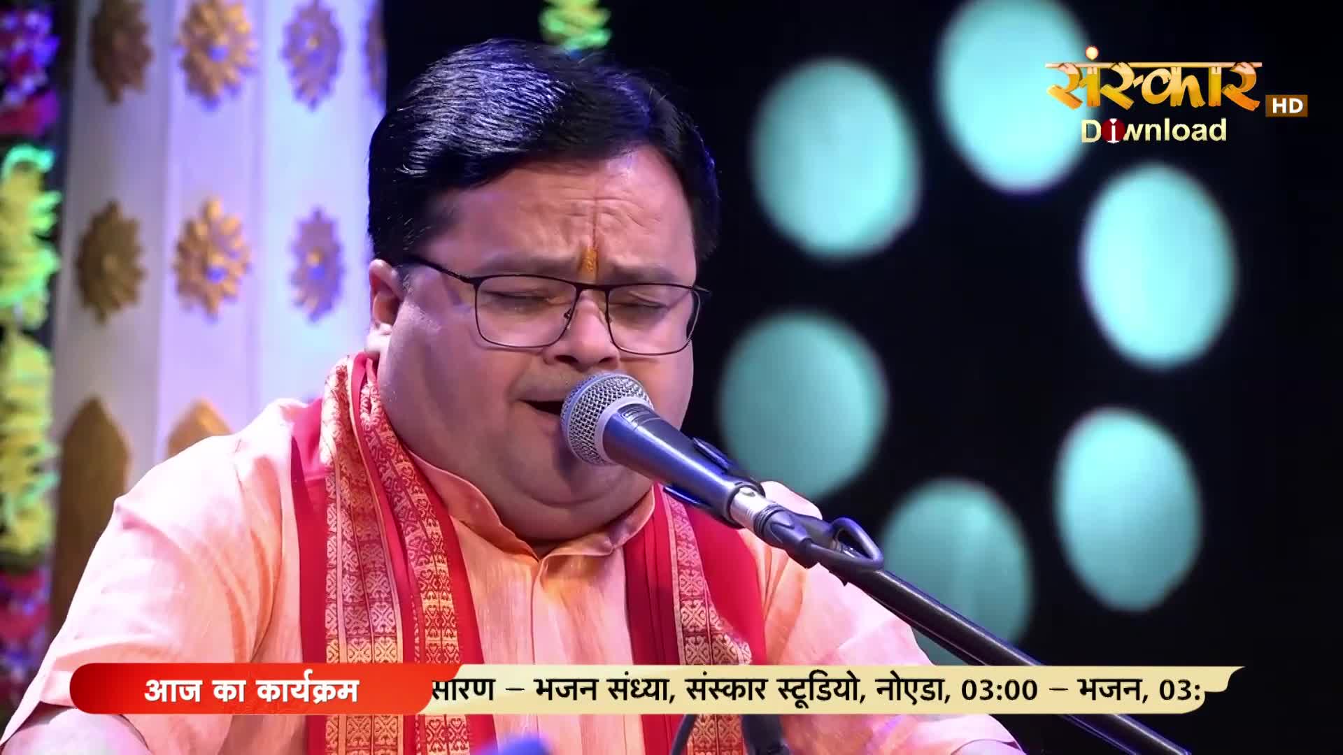 A man in a peach-colored kurta and red scarf sings into a microphone, his eyes closed in concentration. The Sanskar TV logo is visible in the upper right corner.