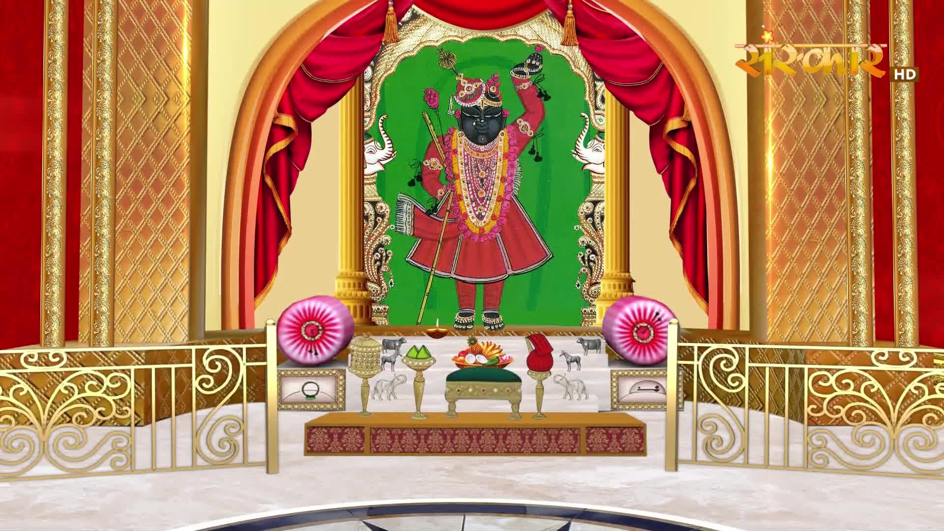 A vibrant depiction of a deity stands on a raised platform, adorned with garlands and a red robe. Ornate gold railings frame the scene, with plush pink cushions on either side.