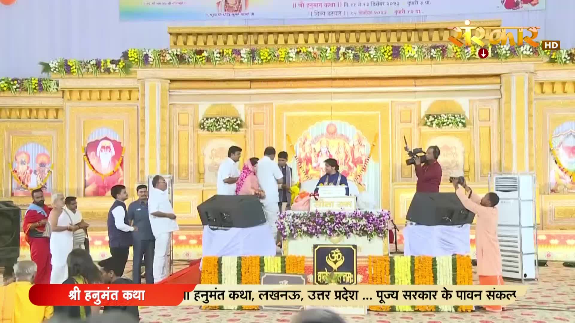 A man in an orange robe holds a camera steady, filming the stage where a speaker sits behind a podium. Another cameraman, dressed in maroon, adjusts his equipment to capture the scene.