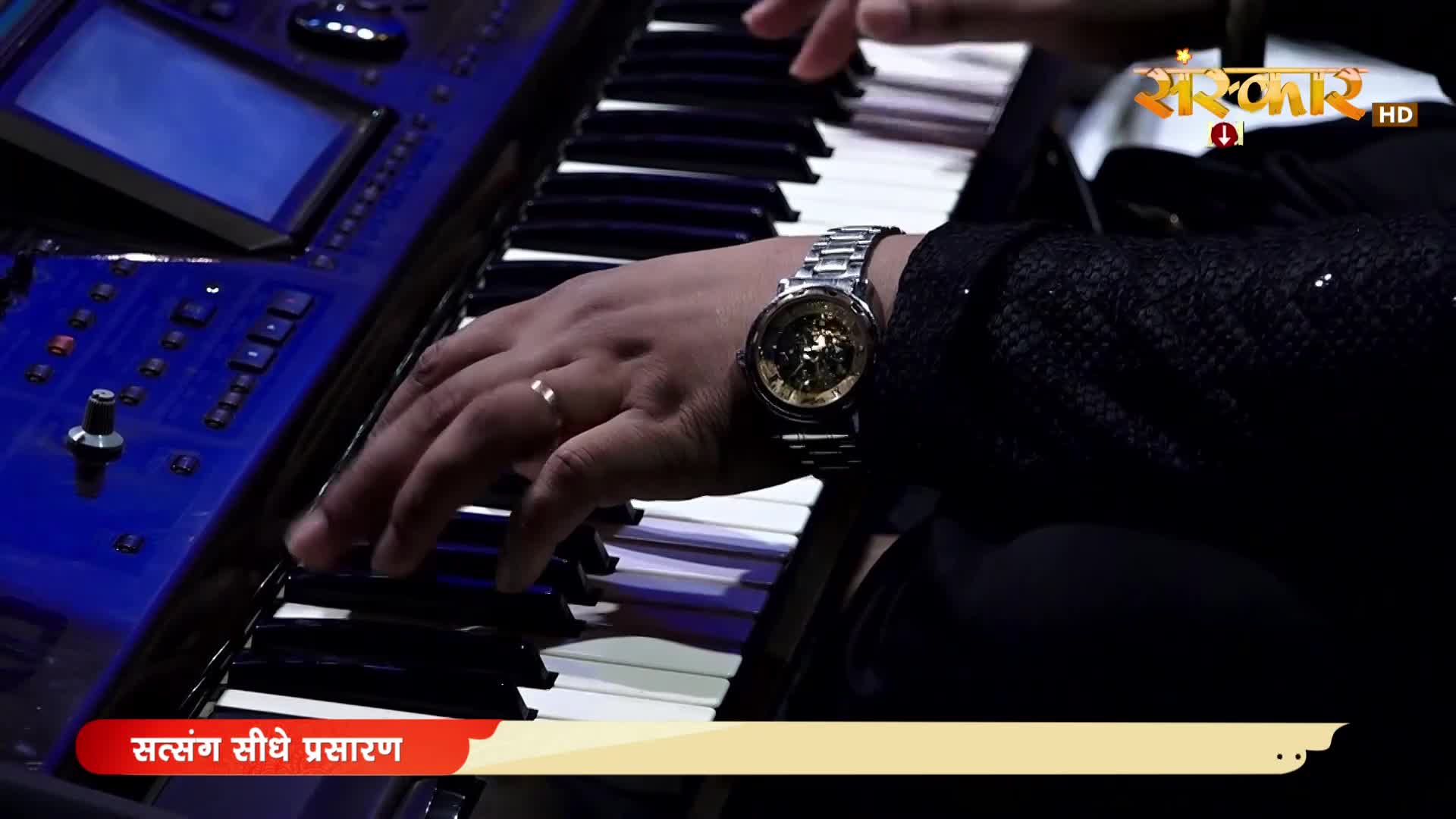 Fingers dance across the keys of a blue synthesizer, creating music for the Sanskar TV broadcast. A gold watch gleams on the player's wrist as the live program continues.