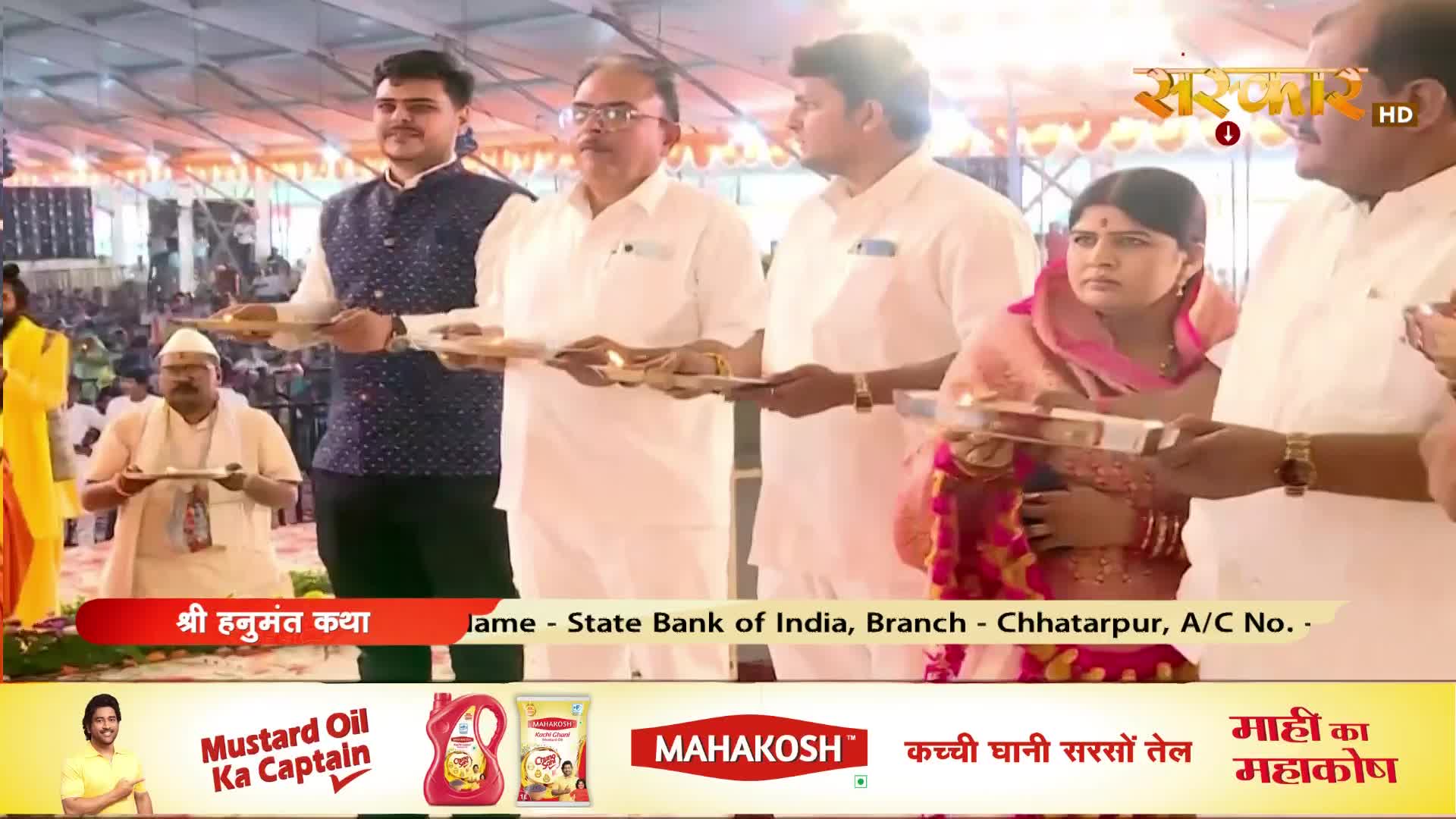 A group of people in white attire stand holding offerings as part of a ceremony. The Sanskar TV logo appears in the upper right corner.
