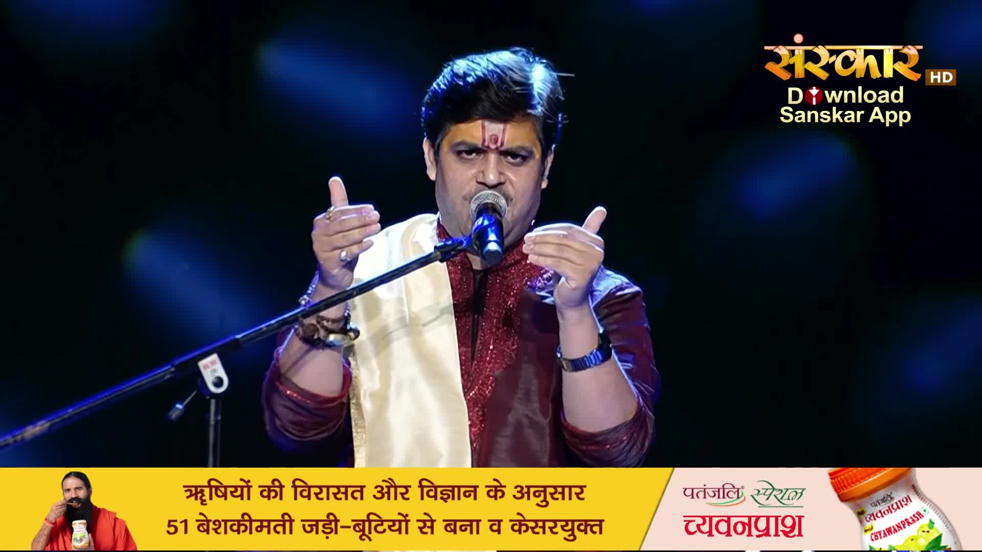 A man in a maroon and gold outfit sings into a microphone, his hands gesturing. A banner below advertises Patanjali Chyawanprash.