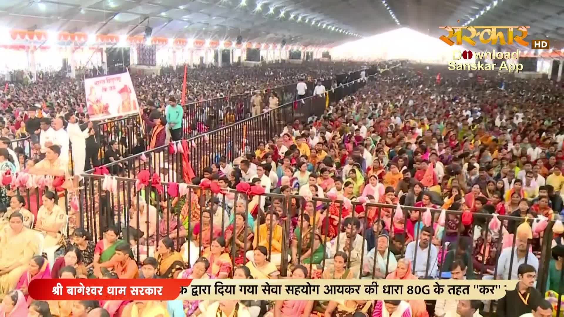 Thousands of people fill a vast tent for a gathering, their colorful clothing a vibrant tapestry under the bright lights. A large banner featuring a figure in saffron is held aloft near the front, drawing attention from the seated crowd.