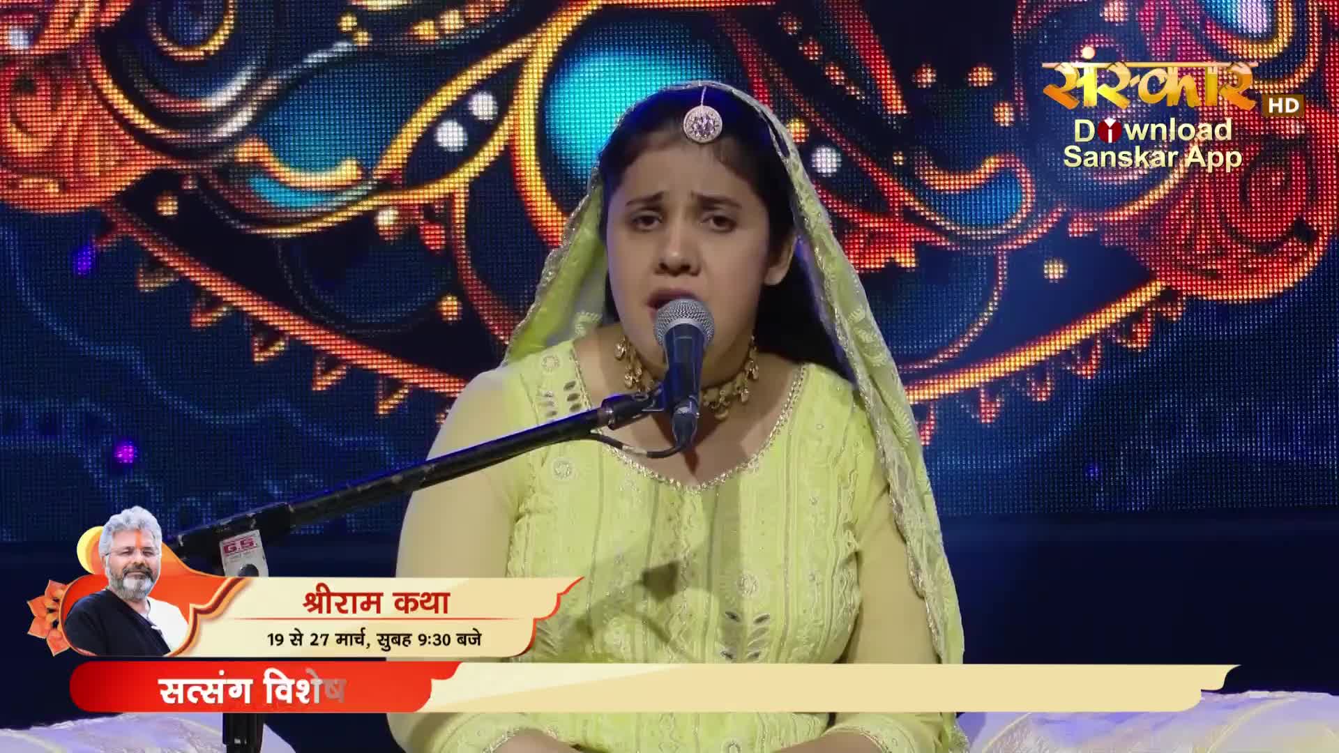 A woman in a light green sari sings into a microphone, her eyes closed. Behind her, a vibrant screen displays intricate patterns and the Sanskar TV logo.