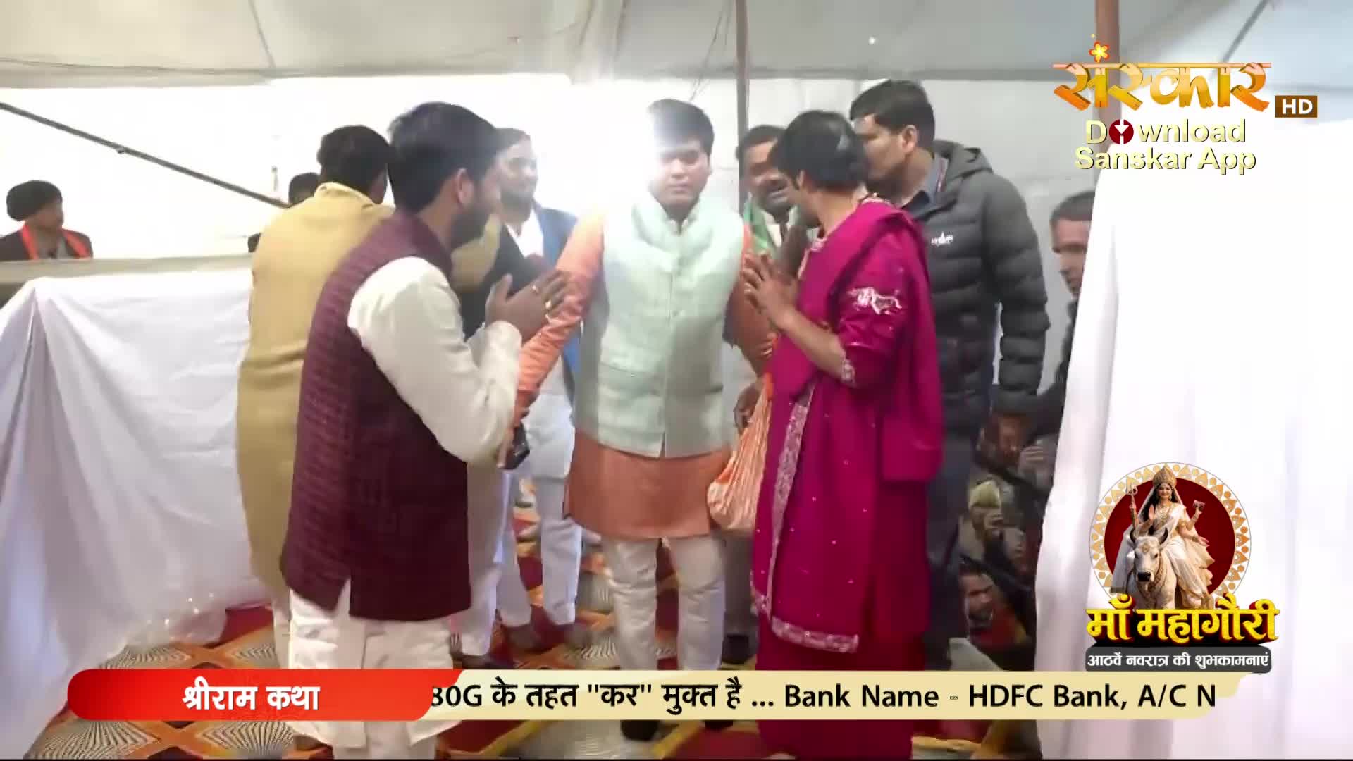 A man in a maroon vest and white kurta extends his hands, palms together, towards another man in a pale green vest over an orange kurta. A woman in a vibrant pink outfit stands to the right, facing the man in the orange kurta.