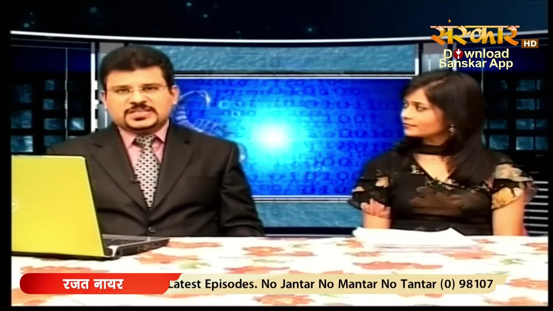 A man in a suit and tie sits at a desk, a laptop open before him. Beside him, a woman in a patterned top looks off-camera. The Sanskar HD logo appears in the upper right corner.