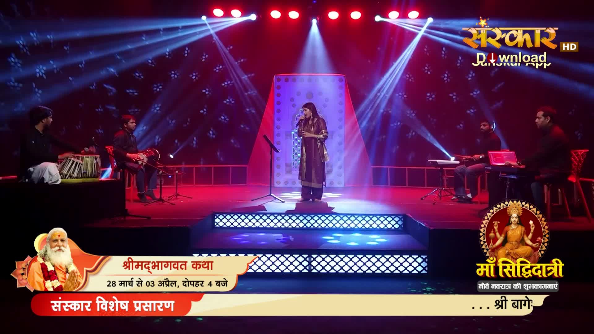A woman stands on a raised platform, singing into a microphone, bathed in dramatic stage lights. To her left, a musician plays the tabla, while to her right, another plays a keyboard.