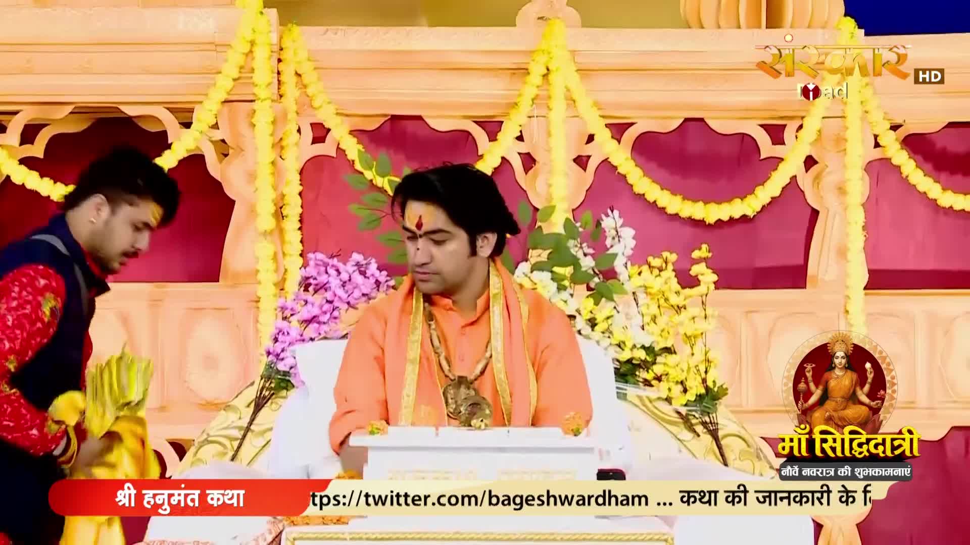 A man in an orange robe sits on a white platform, looking down. Another man, dressed in a patterned vest over a red shirt, approaches the platform, holding yellow fabric.